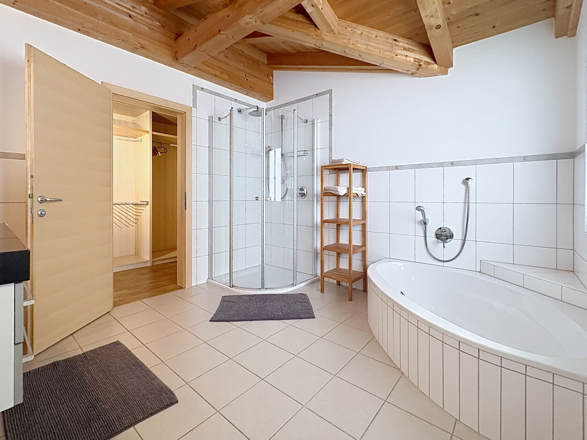 A bathroom with a glass shower enclosure, a wooden shelf, a bathtub, and a door leading to a walk-in closet.