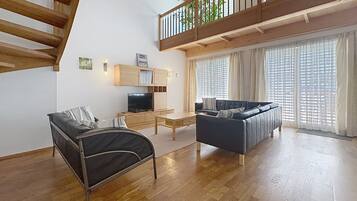 Ein modernes Wohnzimmer mit einer Couch, einem schwarzen Sessel, einem Fernseher, Holzböden und einem Balkon mit Pflanzen.