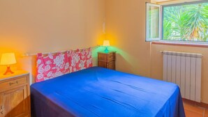 Un dormitorio con cama, mesita de noche, lámpara, ventana y vistas a zonas verdes, y un radiador.