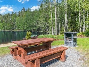 Una mesa de picnic con bancos, un basurero y un pequeño hogar para fuego en un parque con un lago y árboles.