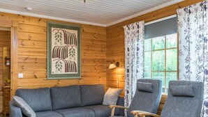 En mysig stuga med träpanel på väggarna, en grå soffa, två fåtöljer, ett litet bord med en blomma och ett fönster med blommiga gardiner.