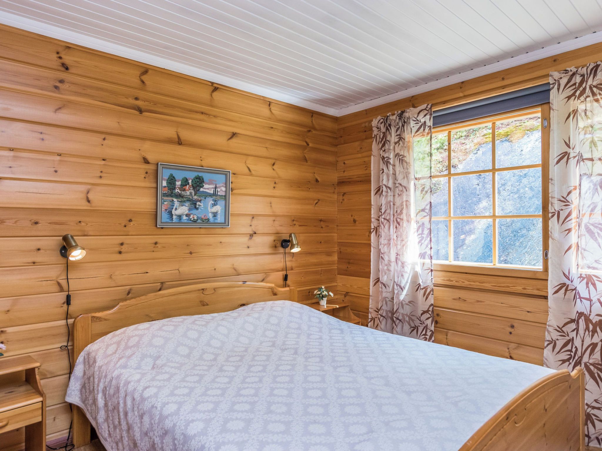 Een slaapkamer in een houten chalet met een bed, twee bedlampen, een lijst met een afbeelding aan de muur, en een raam met gordijnen.