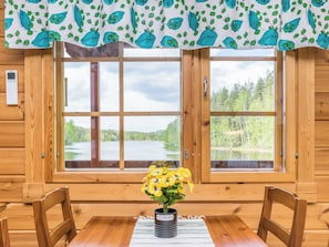A wooden cabin interior with a large window overlooking a lake, a table with chairs, and a potted plant on the windowsill.