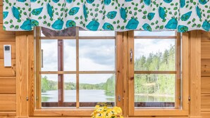 En stuga av trä med ett stort fönster med utsikt över en sjö, ett träbord med stolar och en krukväxt med gula blommor.