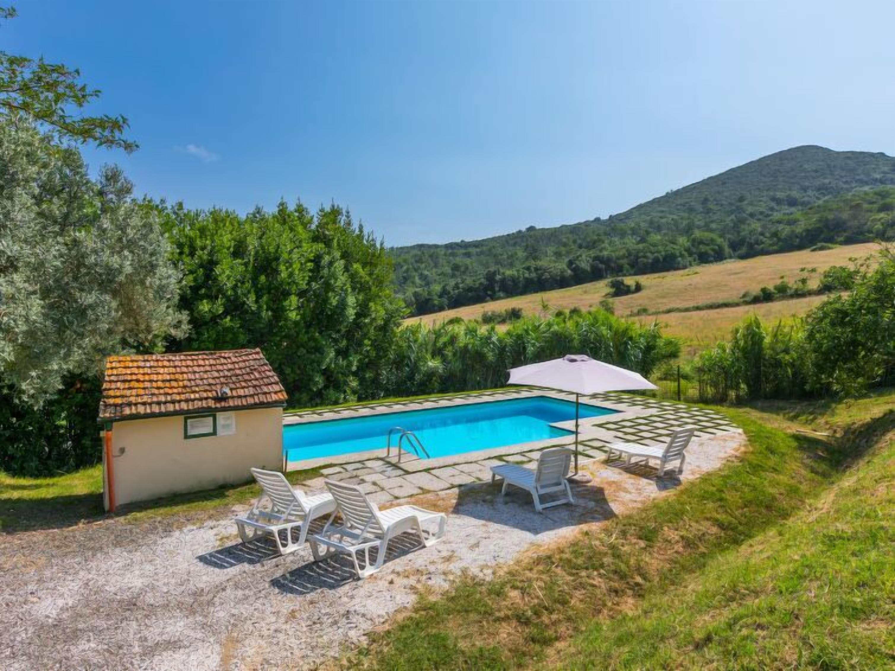 A swimming pool with a small adjacent building, surrounded by outdoor seating and a view of a hilly landscape.