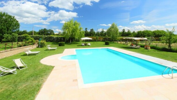 Ein rechteckiger Pool mit geschwungener Kante, umgeben von einer gepflasterten Liegefläche. Im Hintergrund sind Liegestühle und Sonnenschirme zu sehen.