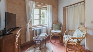A cozy room with a wooden ceiling, a TV on a wooden cabinet, wicker chairs, a small round table, and a window with curtains. - Casa di Nena by Interhome (Camaiore)
