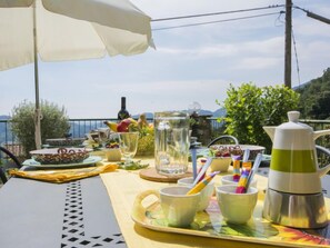 Een tafel buiten, met uitzicht op zee, een witte parasol en diverse serviesartikelen.