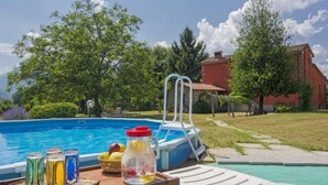 A swimming pool with a sun lounger, a table with drinks and fruit, and a red building in the background. - San Bernardino by Interhome (Barga)