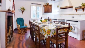A dining area with a wooden table set for four, surrounded by wooden chairs. A blue upholstered chair is positioned to the left. The room features exposed wooden beams on the ceiling, a window with white shutters, and a kitchen area with white cabinets and a sink. - San Bernardino by Interhome (Barga)