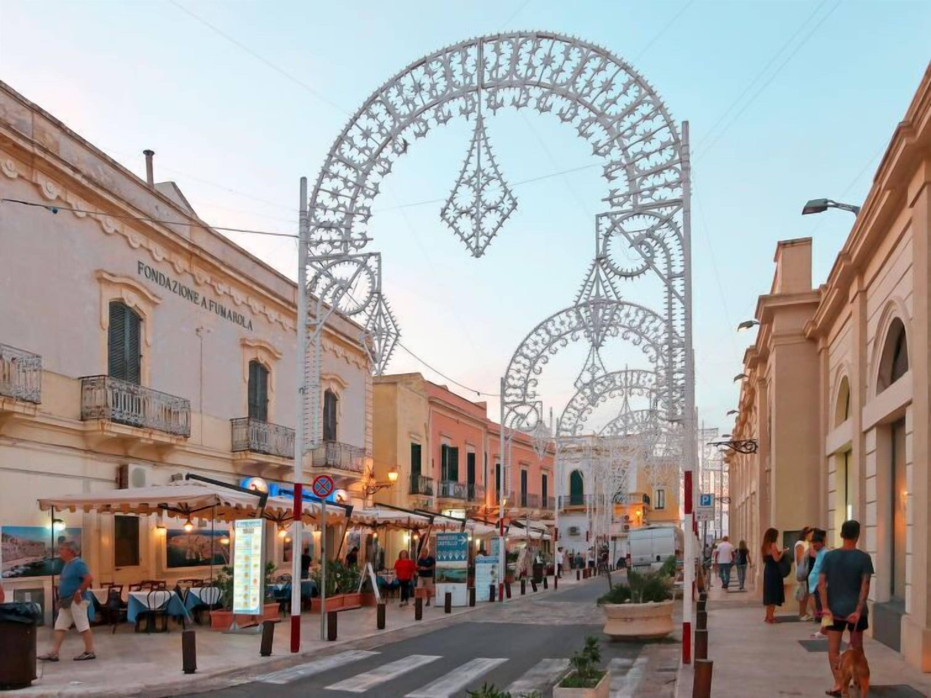 Une rue bordée de grilles en métal ouvragées, un restaurant avec une terrasse, et un bâtiment portant l’inscription « FONDAZIONE A FUMAROLA ».