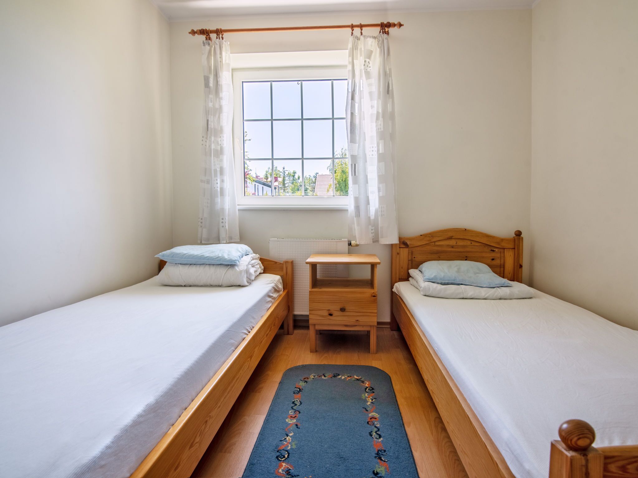 A small, clean room with two single beds, a wooden nightstand, a small table, and a window with curtains.