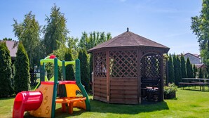 A playground with a slide and climbing structure, a wooden gazebo, and a picnic table in a grassy area with trees in the background. - Mały dworek by Interhome (Leba)