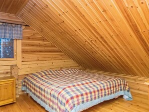 Una stanza con soffitto spiovente, pareti in legno, un letto con copripiumino a quadretti, un comodino in legno e una finestra con tenda.