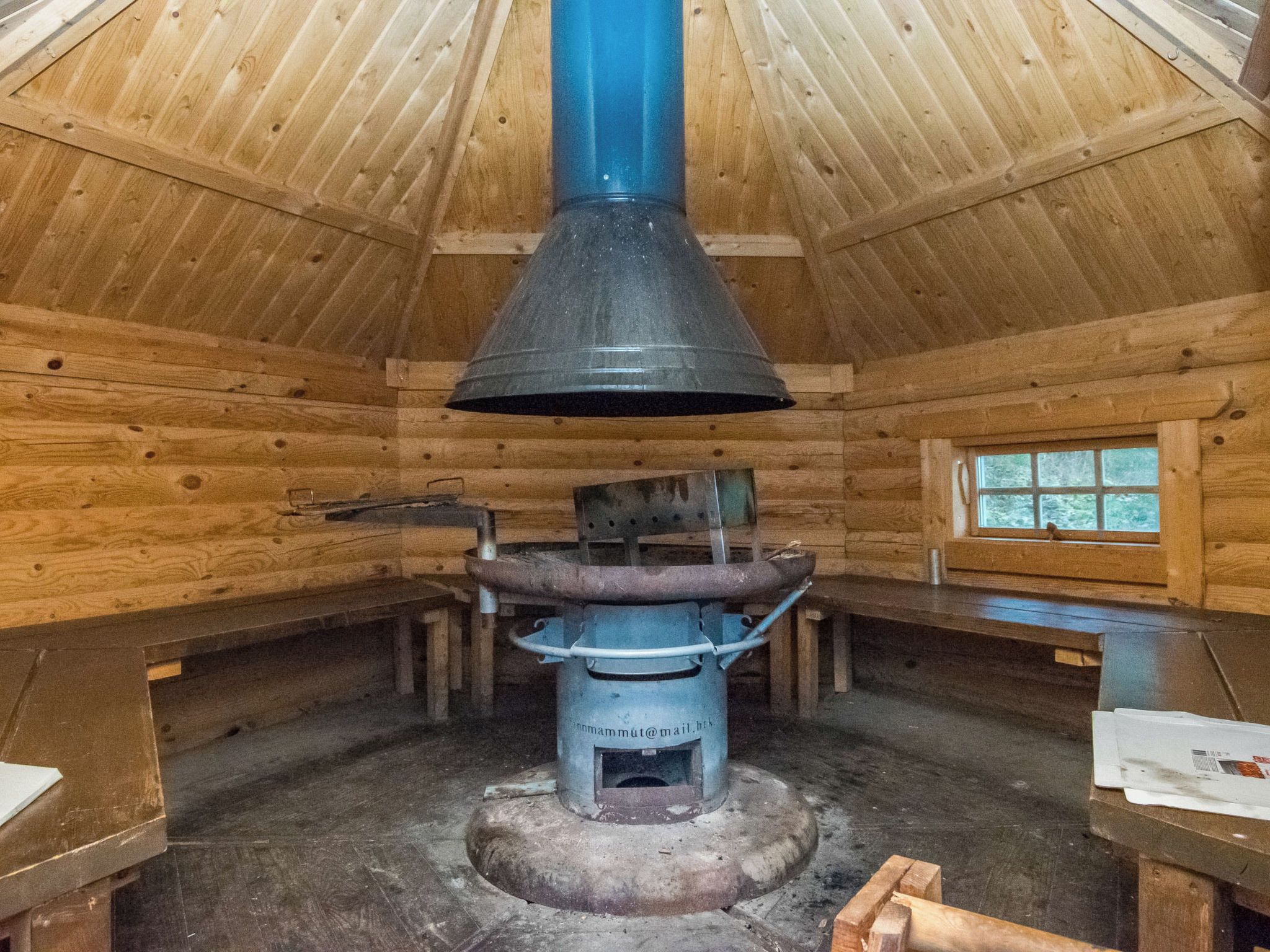 A wooden cabin interior with a central wood stove, benches, and a window.