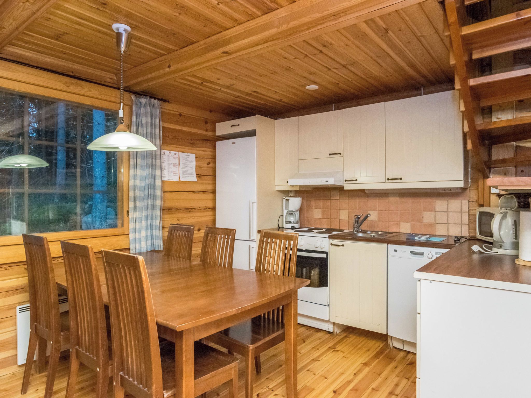En inredning med träpanel, ett matbord, stolar, ett kök med vita skåp och ett fönster med gardiner.
