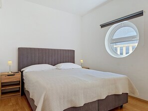 A bedroom with a bed, bedside tables, a round window, and a view of buildings outside. - Apartment Anđa by Interhome (Trogir/Slatine)