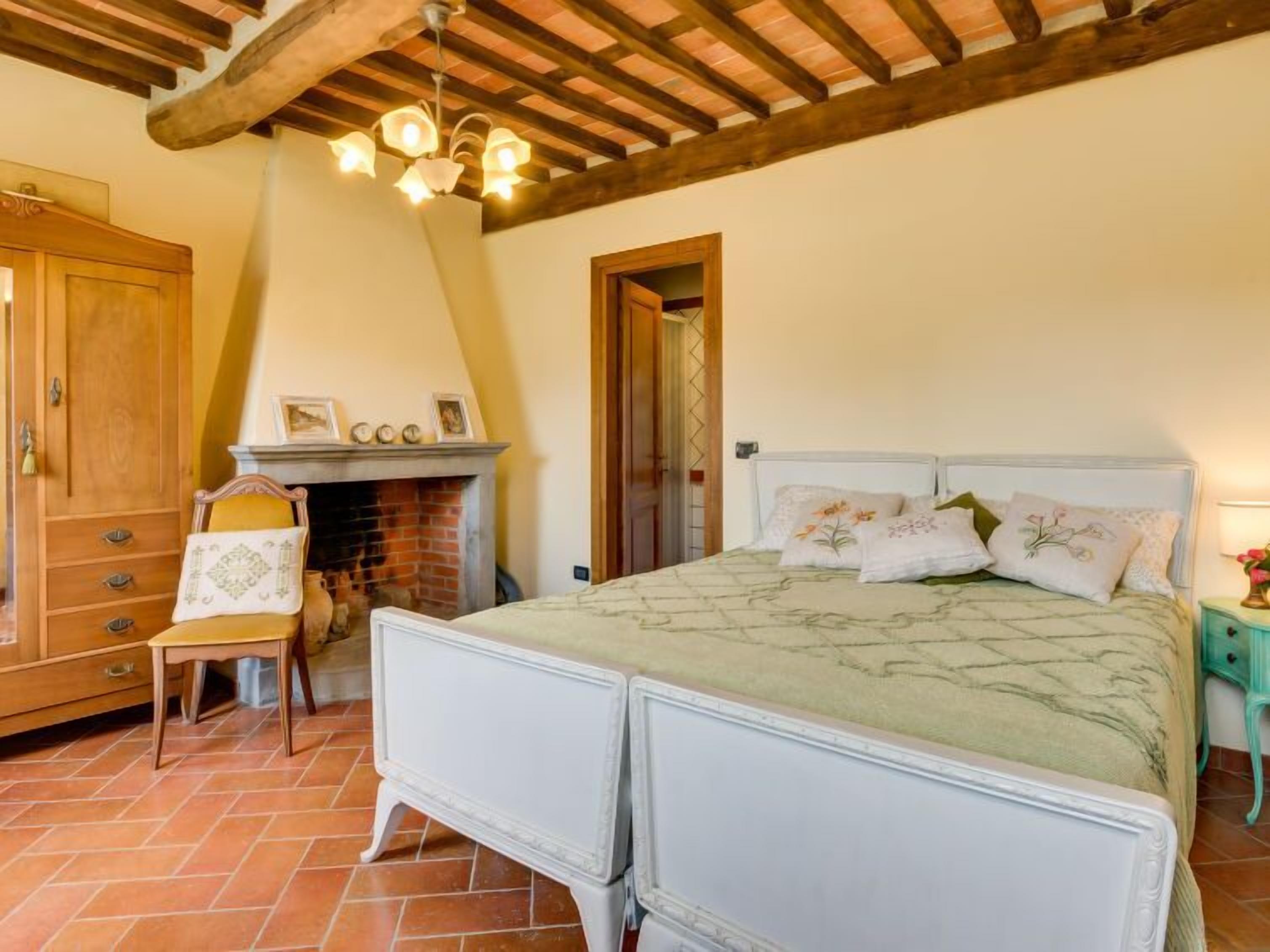 A bedroom with a bed, a wooden wardrobe, a chair, a fireplace, and a wooden ceiling.