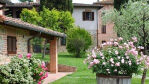 Un bâtiment en pierre avec un toit en tuiles, un chemin en briques et un grand bac à fleurs en bois, orné de fleurs roses.