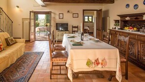 Un espace repas avec une longue table dressée pour six personnes, des chaises en bois, un téléviseur et une vue sur l’extérieur.