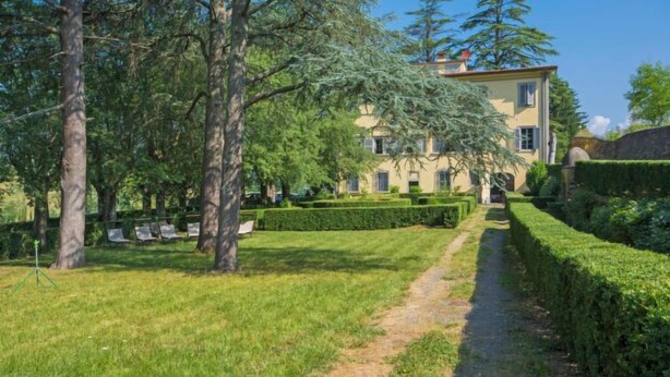 Ein gepflegter Garten mit einem Weg, der zu einem großen, klassizistischen Gebäude mit zahlreichen Fenstern und einer symmetrischen Fassade führt.