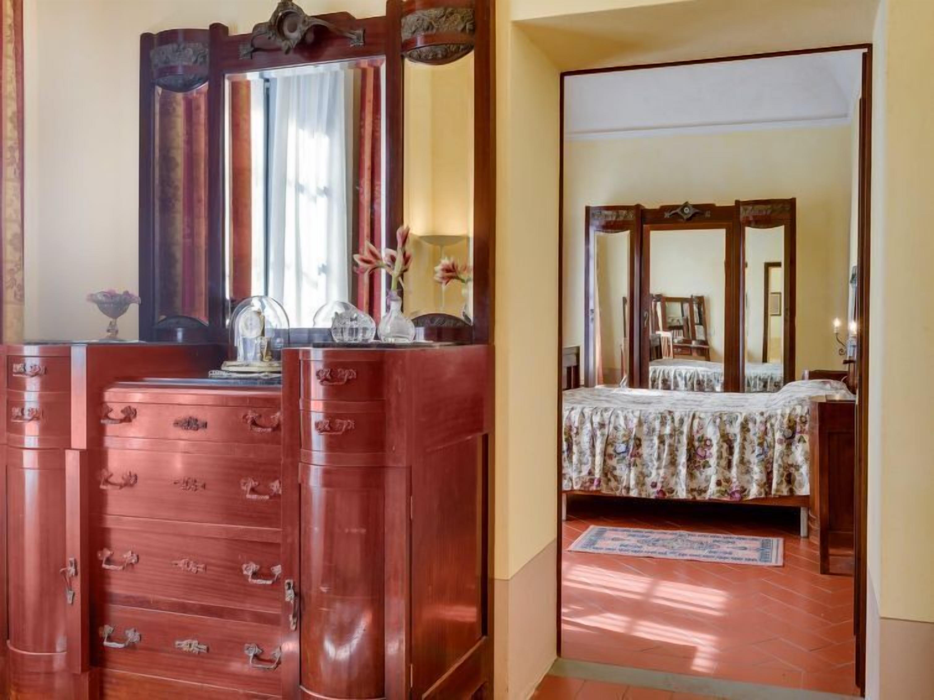 Une pièce comprenant une commode en bois, un grand miroir et un lit recouvert d’une couvre-lit à motifs floraux.