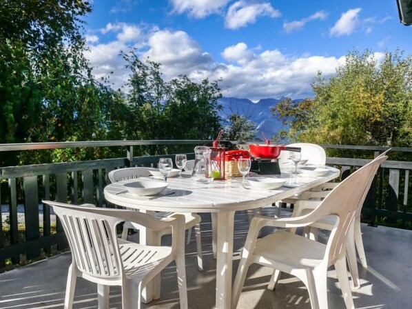 A round outdoor table with white chairs, a red pot, and a grill, set against a scenic mountain backdrop.