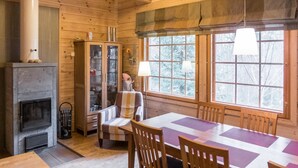 A wooden cabin interior with a fireplace, a dining table with chairs, and large windows.