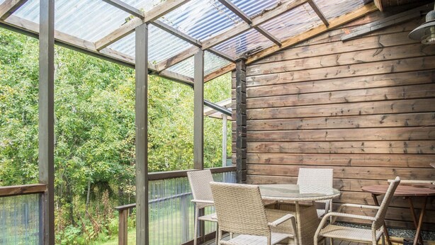 A covered patio with wooden flooring and wicker furniture, overlooking a garden.