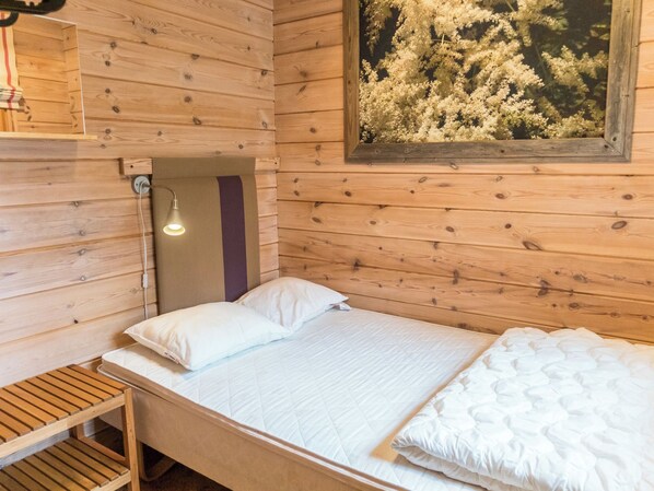A single bed with white bedding in a wooden cabin room with a wall-mounted light fixture and a framed picture of flowers.