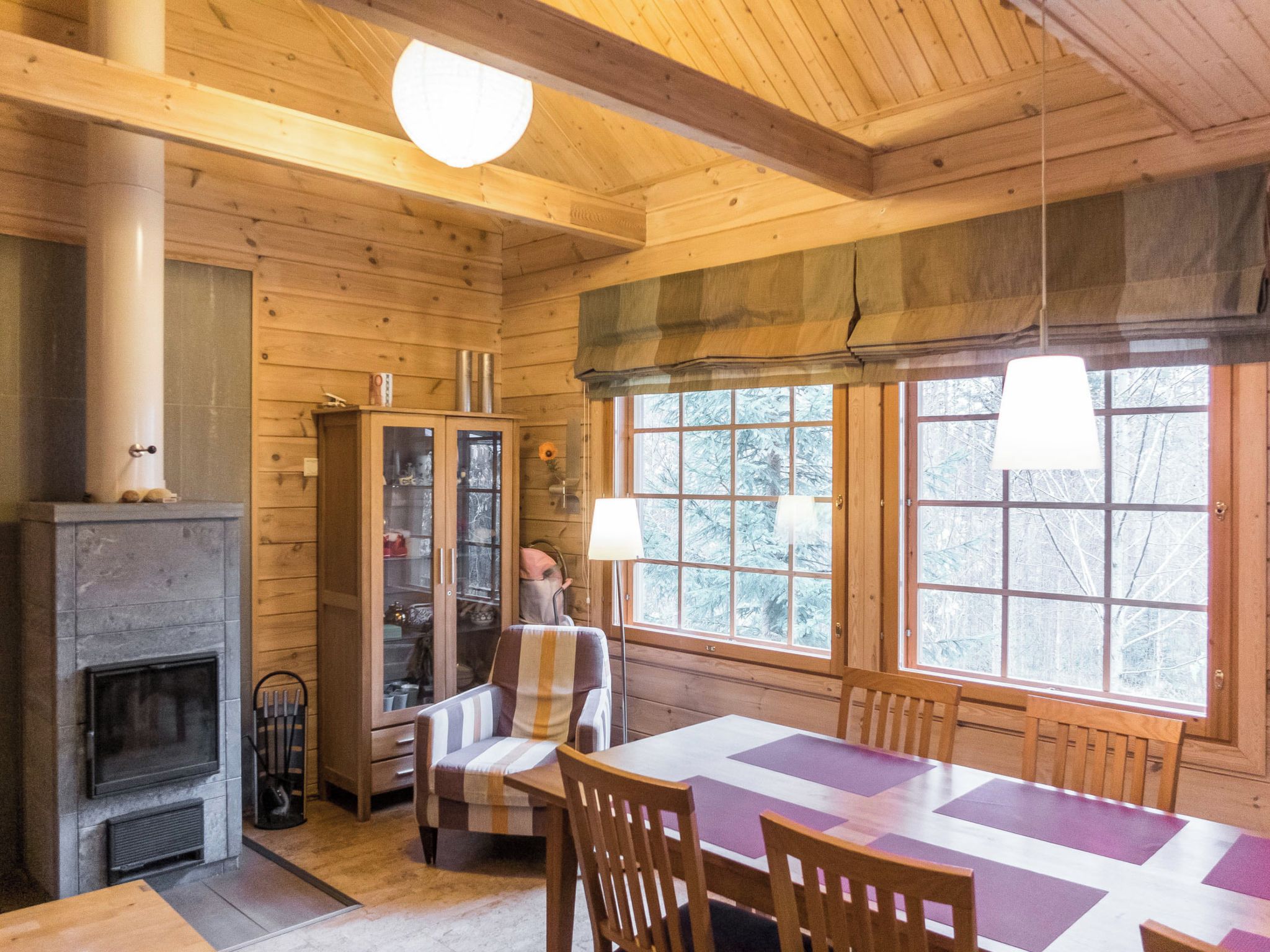 A cozy cabin interior with a fireplace, wooden walls, a dining table, and large windows.