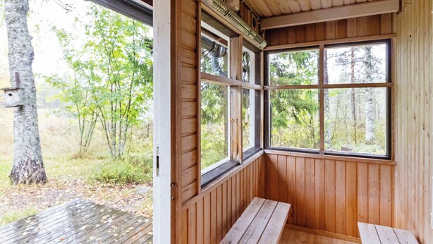 A wooden cabin with a bench and a table outside, a window with a view of trees, and a door.
