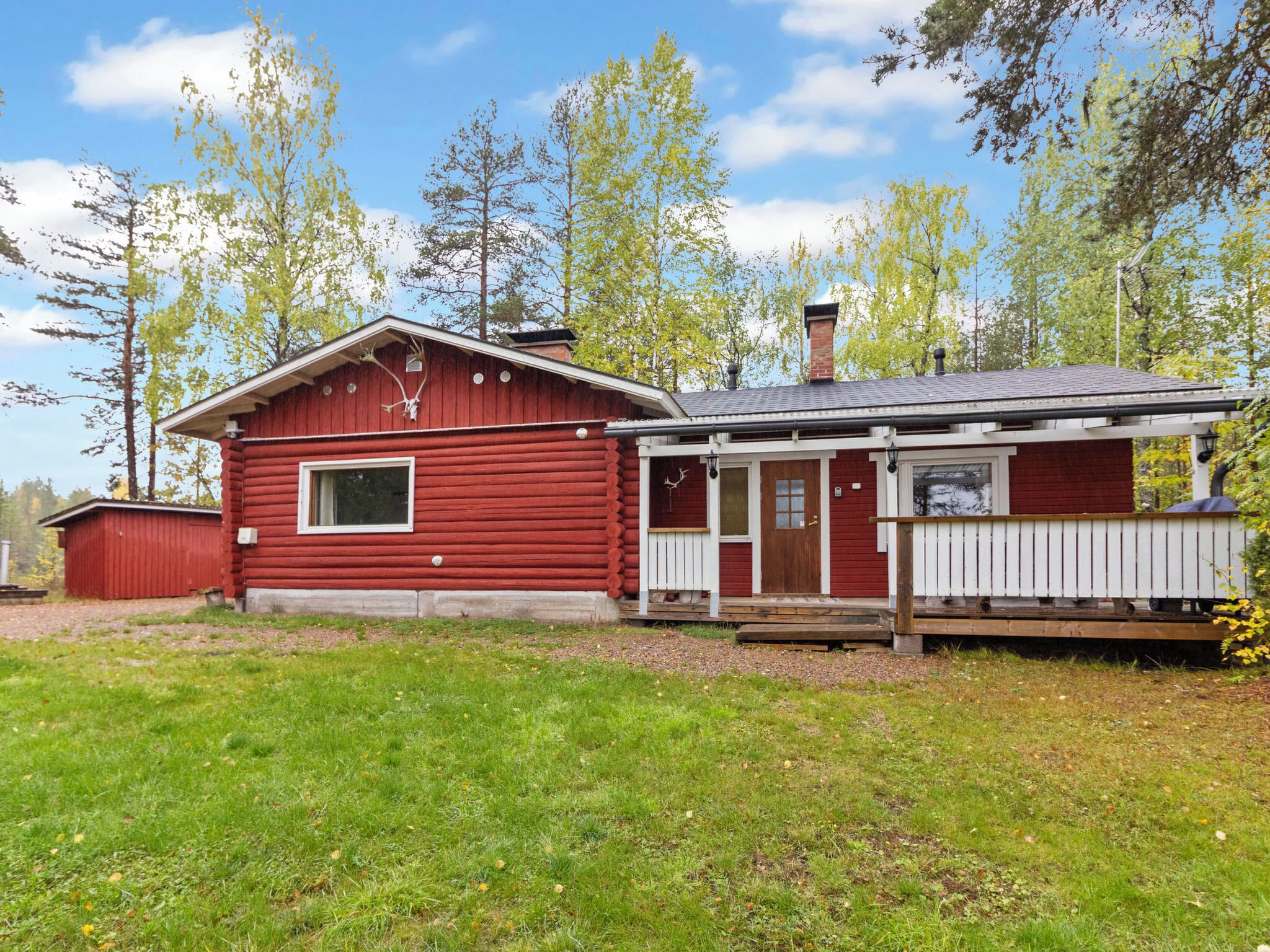 A red log cabin with a porch and a small shed in a wooded area.