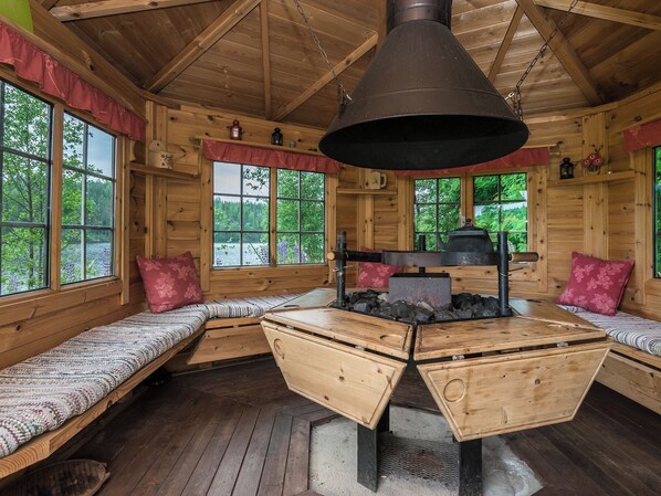 Interior de una cabaña con paredes de madera, amplias ventanas, una chimenea central y zonas de estar.