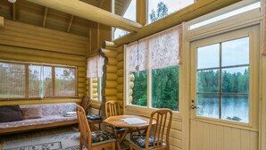 Interior acogedor de una cabaña con pared de madera, zona de comedor con sillas y mesa, banco con cojines y ventanas amplias con vistas a un lago.