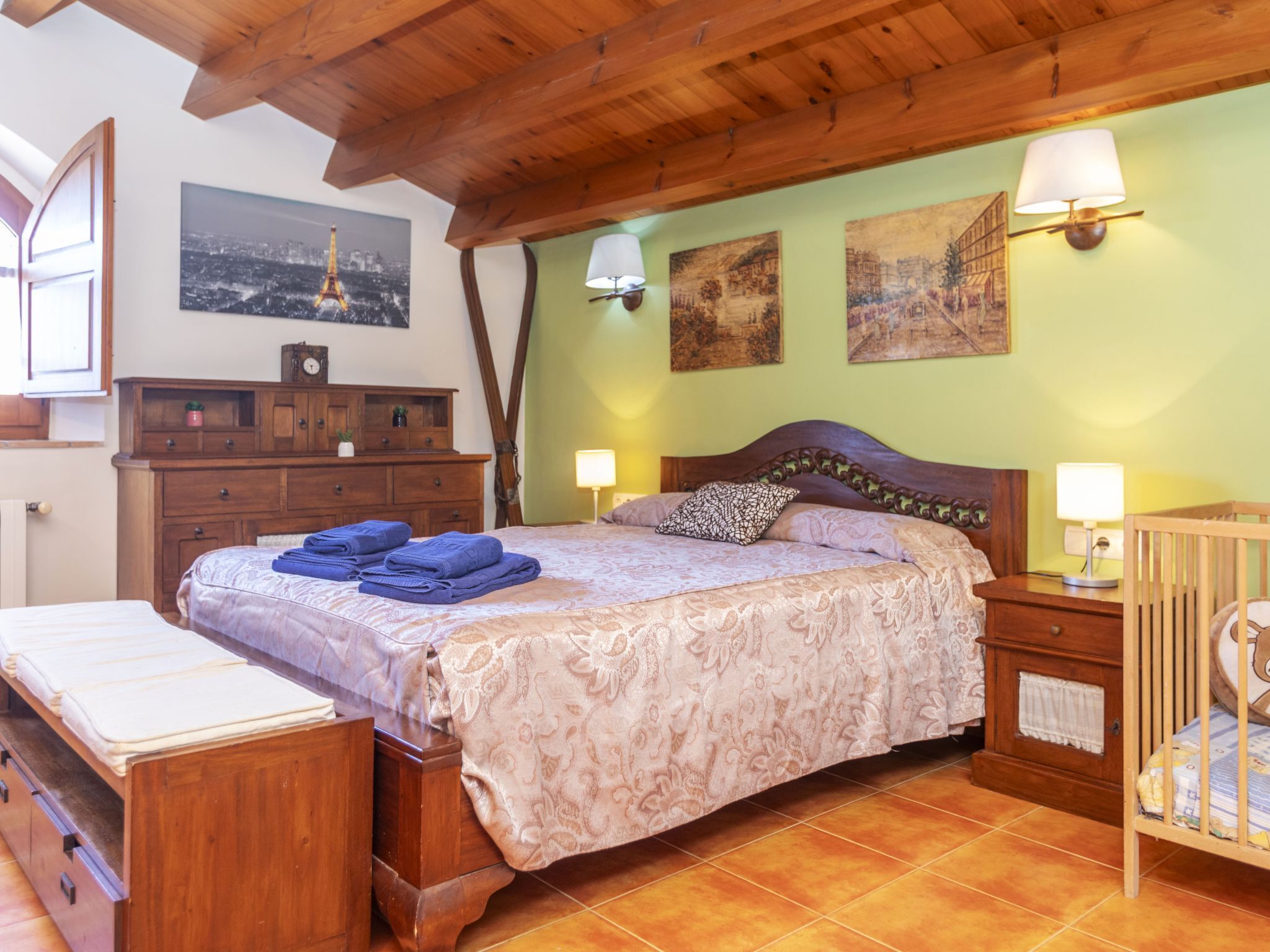 A bedroom with a wooden bed, two bedside lamps, a crib, a dresser, and artwork on the walls.