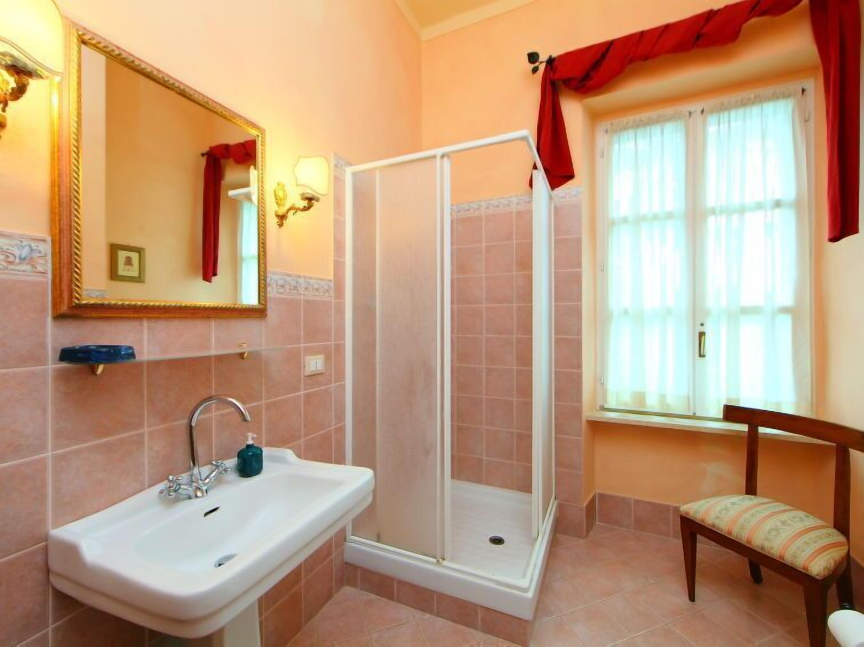 Un baño con lavamanos, cabina de ducha, una silla y ventana con cortinas.