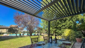 Zona d'esbarjo exterior amb taula de vidre preparada per a quatre persones, amb caires i sota una pérgola.