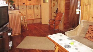 A cozy room with a wooden interior, a sofa, a table with a patterned tablecloth, a TV, and a bar area with stools.