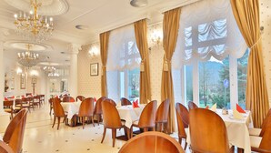 A dining area with multiple tables and chairs, large windows with curtains, and chandeliers. - Sun Palace Wellness Hotel Marianske Lazne (Marianske Lazne)