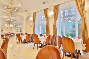 A dining area with multiple tables and chairs, large windows with curtains, and chandeliers. - Sun Palace Wellness Hotel Marianske Lazne (Marianske Lazne)