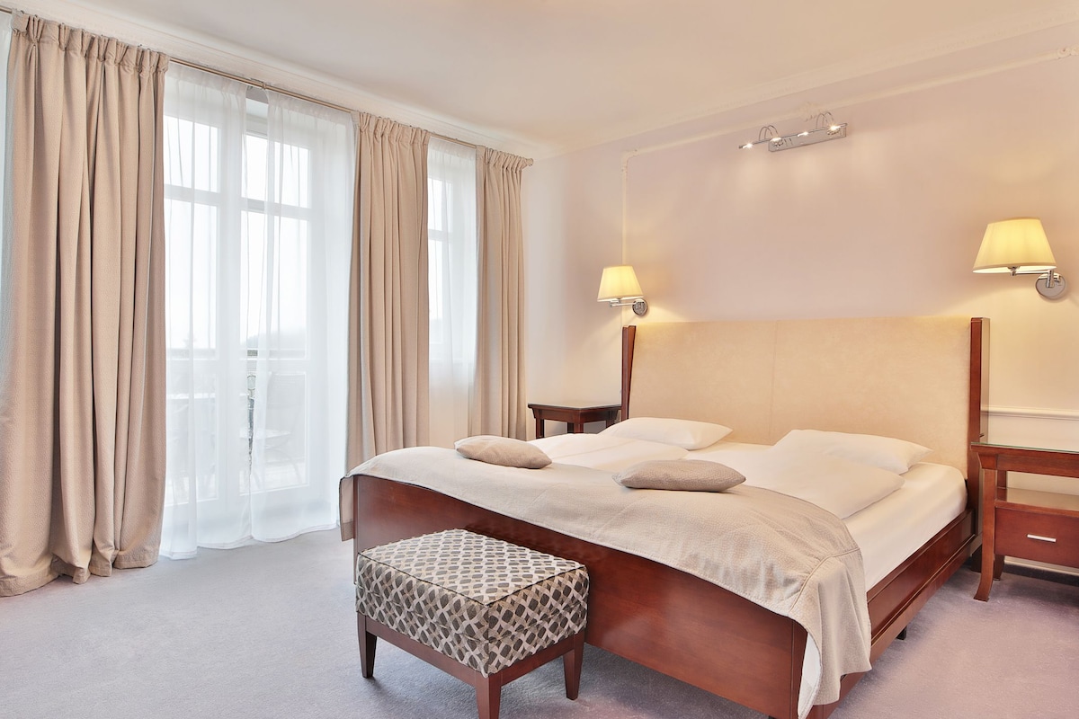 A neatly arranged bedroom with a large bed, two bedside tables, a nightstand, and a patterned ottoman.