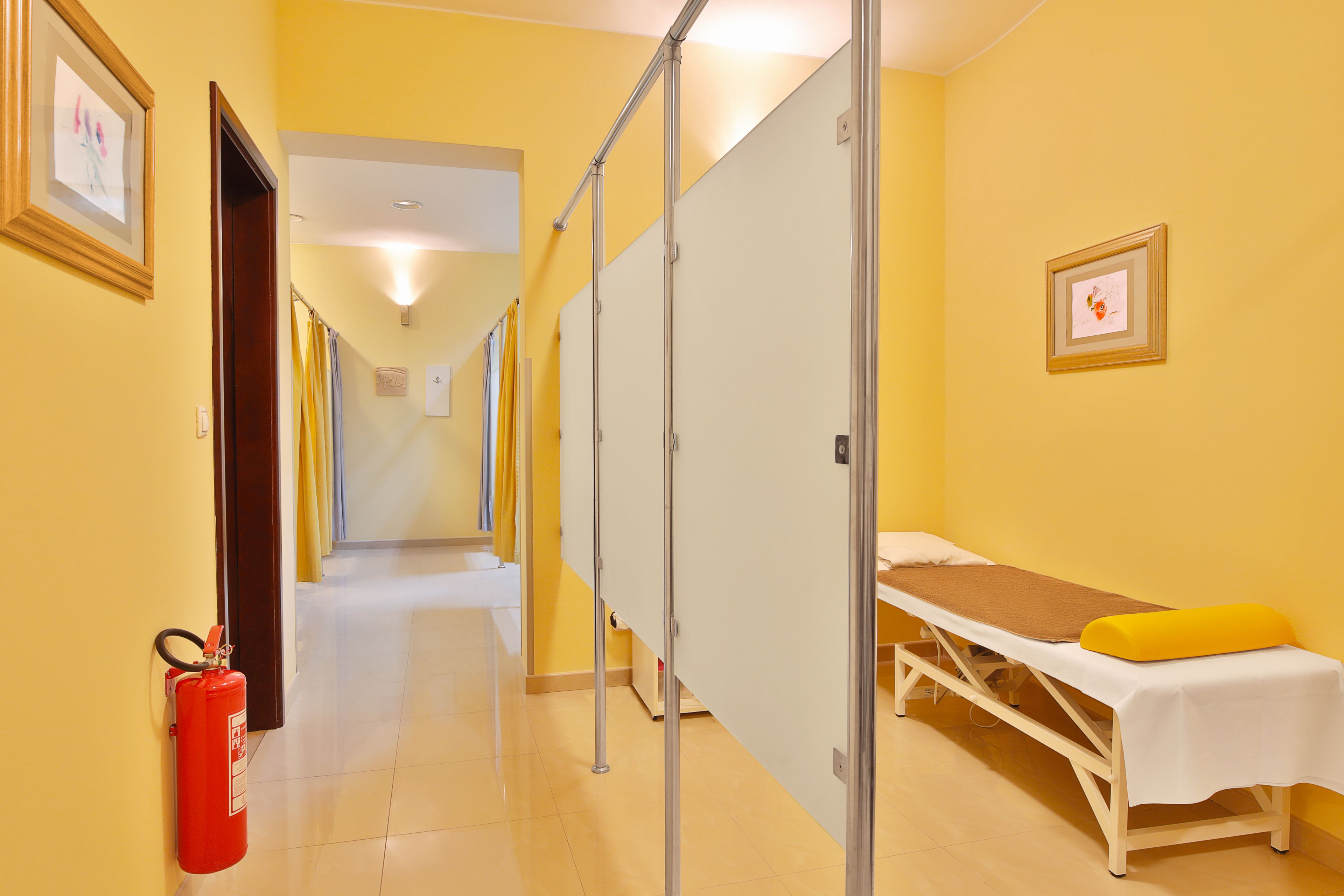 A corridor with a bed, a fire extinguisher, and a framed picture on the wall.