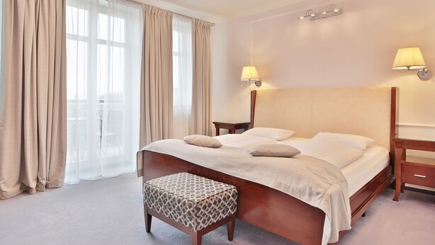 A hotel room with a bed, two bedside tables, a nightstand, a window with curtains, and a wall-mounted lamp. - Sun Palace Wellness Hotel Marianske Lazne (Marianske Lazne)