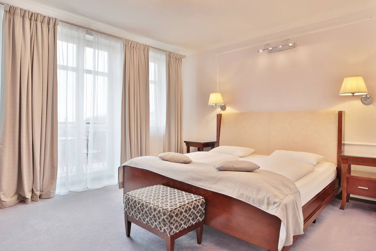 Ein ordentlich eingerichtetes Schlafzimmer mit einem großen Bett, zwei Nachttischen, einem Beistelltisch und einem gemusterten Hocker.