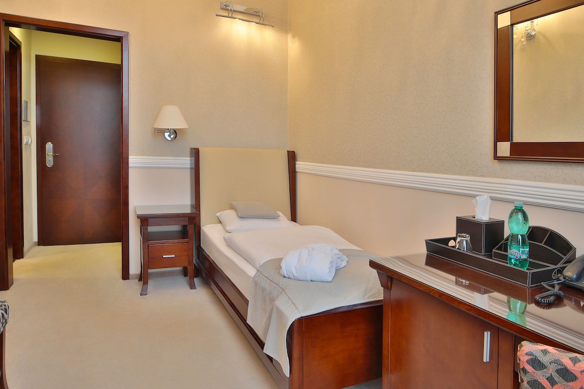 Quarto de hotel com cama, mesa de cabeceira, luminária e escrivaninha com telefone e uma garrafa.