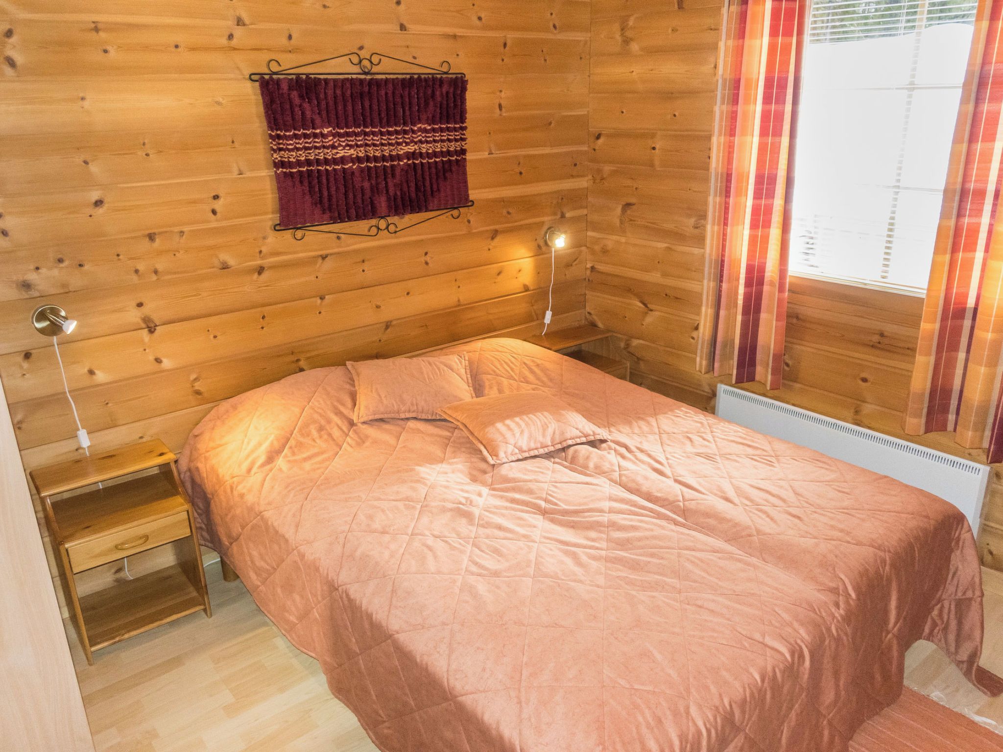 Ein Zimmer mit Holzboden, einem Bett, einem Nachttisch, einer Wandlampe und einem Fenster mit Vorhängen.