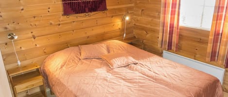 Ein Zimmer mit Holzboden, einem Bett, einem Nachttisch, einer Wandlampe und einem Fenster mit Vorhängen.