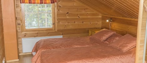 Une petite chambre mansardée, chaleureuse et accueillante, avec un plafond en pente, une fenêtre avec des stores, un lit recouvert d’une courtepointe et une lampe de chevet.