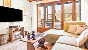 A cozy living room with a wooden ceiling, a large window with a mountain view, a flat-screen TV, and a comfortable sofa.